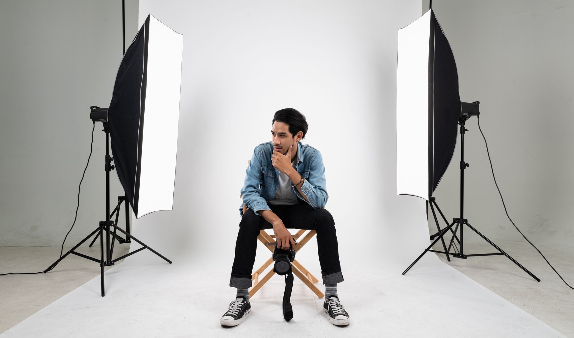 Portrait photo of young smart Asian professional photographer sitting on white photo backdrop indoor studio set with set of strobe flash and softbox preparing for product and model shoot.